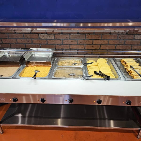 A buffet line with various Mexican dishes, including rice, tortillas, and taquitos, against a brick wall.